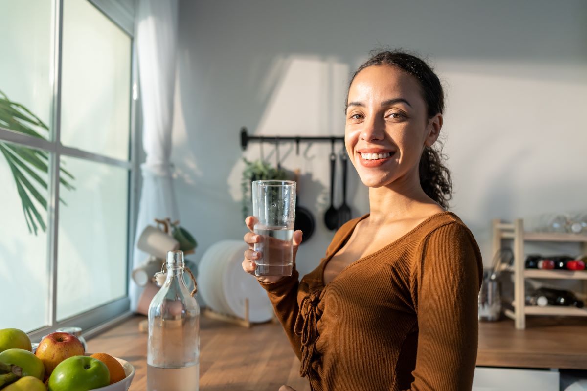 water pitcher sitting on counter next a Women holding a clear glass of purified and filter water close to her face.